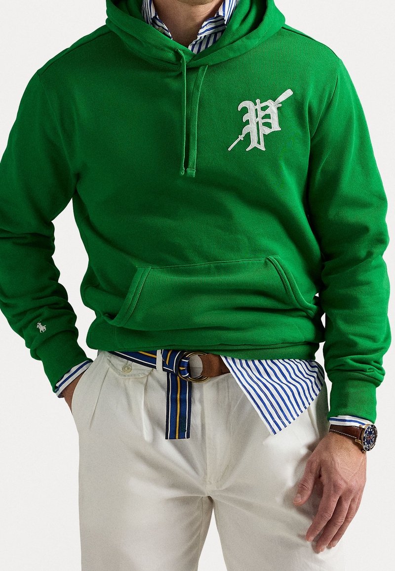 Homme portant un sweat à capuche vert avec un "P" blanc brodé et un pantalon beige, une chemise rayée bleue et blanche, une ceinture rayée bleue et une montre-bracelet.