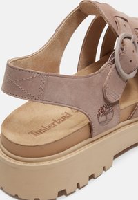 Taupe suède Timberland sandaal met verstelbare gesp en dikke beige profielzool, met geperst merkembleem aan de zijkant.