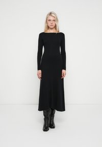 Black ribbed maxi dress with long sleeves, a boat neckline, and a fitted silhouette. Worn with knee-high black boots. Smooth texture.