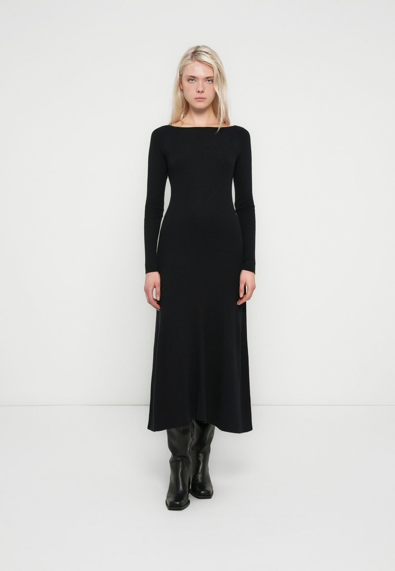 Black ribbed maxi dress with long sleeves, a boat neckline, and a fitted silhouette. Worn with knee-high black boots. Smooth texture.