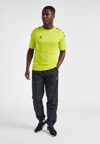 Camiseta atlética verde brillante de manga corta con acentos negros, combinada con pantalones negros y zapatillas negras, todo en un fondo neutro.
