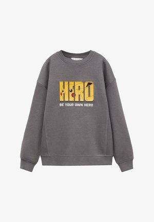 Siv sweatshirt z izrazitim rumenim grafičnim napisom "HERO" z risanim junakom in besedilom "BODI SVOJ JUNAK". Mehak material in sproščen kroj.