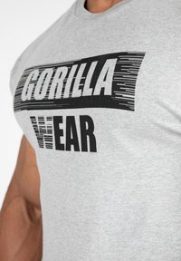 Grijze katoenen T-shirt met korte mouwen. Voorzien van vetgedrukte zwarte tekst met "GORILLA WEAR" in een gestileerd lettertype op de borst.