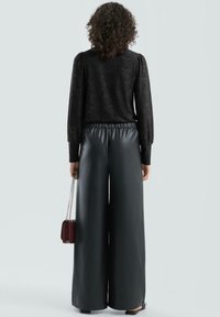Czarna teksturowana bluzka z długim rękawem i bufkowatymi ramionami, zestawiona z szerokimi spodniami z wysokim stanem wykonanymi z ekologicznej skóry. Mała czerwona torebka na łańcuszku.