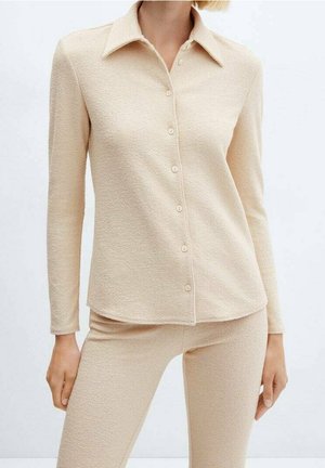 Camicia beige a maniche lunghe con bottoni e colletto, caratterizzata da un tessuto strutturato e orlo arrotondato, abbinata a pantaloni aderenti coordinati.