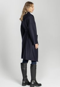 Długi granatowy płaszcz z paskiem w talii, szerokim kołnierzem i pełnymi rękawami. Zestawiony z czarnymi wysokimi botkami do kolan. Gładka tekstura, minimalistyczny design.