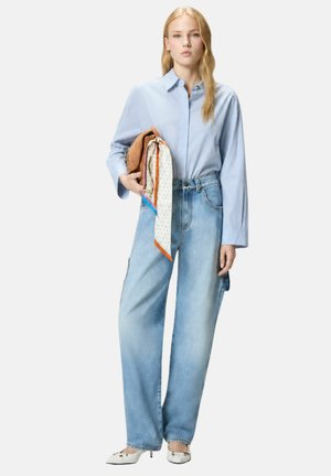 Mujer con camisa azul claro y vaqueros azules holgados, sosteniendo un bolso marrón claro con pañuelo estampado, de pie sobre fondo blanco.