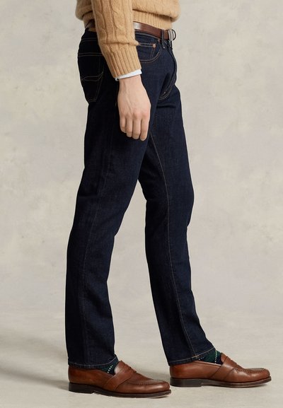 Persona que lleva vaqueros azul oscuro, cinturón de cuero marrón, mocasines de cuero marrón, calcetines verde oscuro y un suéter de punto trenzado color beige.
