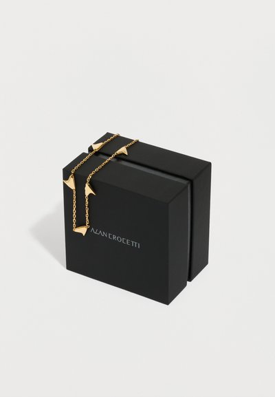 Zelta ķēdes kaklarota ar punktveida bultu formas kuloniem, kas pārklāts pār melnu taisnstūrveida kastīti baltā fona.