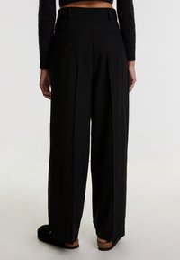 Schwarze weit geschnittene Hose mit hoher Taille, ausgestattet mit Falten, zwei Gesäßtaschen und einem eleganten Finish. Getragen mit schwarzen Clogs.