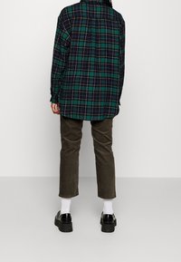 Chemise en flanelle à carreaux verts et bleu marine avec manches longues, détail sur la couture arrière, associée à un pantalon olive et des chaussures noires épaisses. Chaussettes blanches visibles.