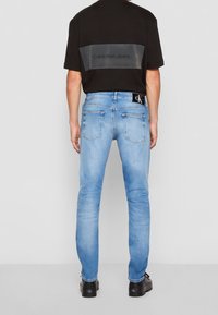 Black T-shirt with a transparent gray panel featuring "Calvin Klein Jeans" text, paired with light blue denim jeans and black shoes.