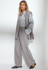 Gilet grigio su misura e pantaloni a gamba larga abbinati a una camicia a righe; tessuto strutturato con una leggera lucentezza; scarpe nere senza lacci base.