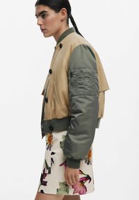 Tan en groene bomberjack met een ritssluiting, grote knopen en een patchzak op de arm, gecombineerd met een rok met bloemenprint.