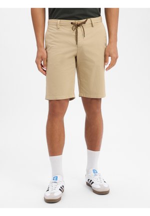 Mann trägt beige knielange Shorts mit braunem Kordelzug, weiße Wadenhochsocken und weiße Adidas-Sneaker mit schwarzen Streifen.