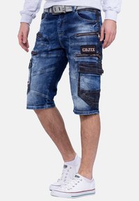 Des shorts cargo en denim bleu foncé dotés de plusieurs poches zippées, de patches décolorés et de coutures contrastées. Comprend une ceinture avec une boucle grise.