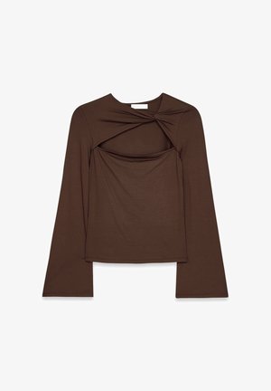 Braunfarbenes langärmliges Oberteil mit tailliertem Design und glockenförmigen Ärmeln, kombiniert mit weißen Hosen. Glatte Textur, minimalistischer Stil.