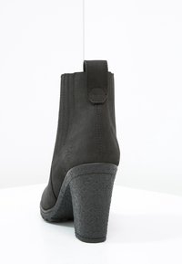 Botte de cheville noire en suede, avec un haut talon texturé, bout rond, panneaux latéraux élastiques et une languette à l'arrière.