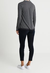 Kvinna stående med ryggen mot kameran, iklädd en grå långärmad tröja, svarta tajta jeans och vita sneakers mot en enfärgad bakgrund.