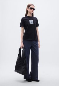 Μαύρο μπλουζάκι με λογότυπο, μπλε ναυτικό παντελόνι με φαρδιά μπατζάκια και μεγάλη μαύρη δερμάτινη τσάντα. Τα γυαλιά ηλίου προσθέτουν μια σύγχρονη πινελιά. Απαλές υφές παντού.