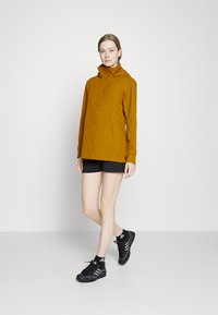 Casaco impermeável amarelo mostarda com capuz, apresentando um fecho de botão, bolsos laterais e um corte descontraído. Calções e sapatos pretos complementam o visual.