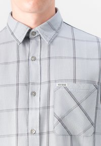 Camisa a cuadros gris claro con cuello abotonado, que presenta un bolsillo con un patrón diagonal y una etiqueta "GUESS" en el bolsillo.