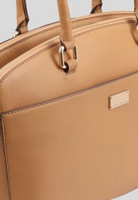 Tan leren handtas met een gestructureerd ontwerp, voorzien van een ritsluiting, goudkleurige hardware en een klein merkembleem aan de zijkant.