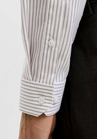 Chemise blanche à rayures brunes étroites. Dotée d'un poignet boutonné avec des boutons blancs et d'une texture lisse. Tatouage visible sur le bras.