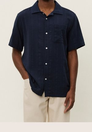 Homme portant une chemise bleu marine foncé à manches courtes avec une poche poitrine, associée à un pantalon beige clair, une main dans la poche.