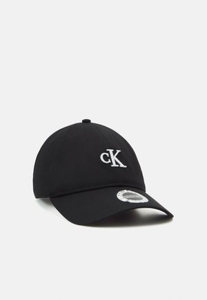 Schwarze Baseballkappe mit weißem "cK"-Logo vorne und einem silbernen Aufkleber auf dem gebogenen Schirm vor einem schlichten weißen Hintergrund.