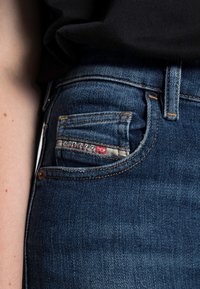 Mörkblå denimjeans med en framficka som har en sydd etikett. Tyget har en slät textur och lätt blekning, med kopparfärgade sömmar som accenter.