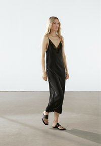 Abito da sera in satin nero con pizzo, caratterizzato da spalline regolabili e un design fluido. Indossato con sandali neri a scivolo su un pavimento neutro.