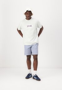 Camiseta verde claro con un gráfico púrpura, combinada con pantalones cortos azul claro. Accesorios incluyen un sombrero de cubo blanco y zapatillas navy.