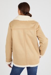 Giacca beige foderata in shearling con un ampio colletto, maniche lunghe e texture morbida. Presenta una forma dritta e dettagli di cucitura visibili.