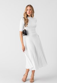 Robe en tricot côtelé blanche avec des manches jusqu'au coude, design de jupe plissée, accompagnée d'un petit sac à main noir et de sandales à talons nudes.