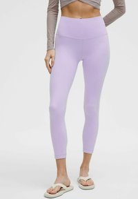 Leggings lilla con vita alta, texture liscia e design aderente. Indossati con infradito chiare.