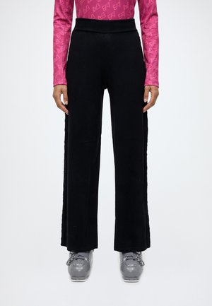 Modèle portant un pantalon noir à jambes larges et à taille haute, un haut rose à motifs et des chaussures de ski grises.