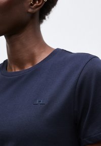 Κοντινό πλάνο ατόμου που φοράει σκούρο μπλε T-shirt με στρογγυλή λαιμόκοψη και μικρό ανάγλυφο λογότυπο στο αριστερό στήθος, και χρυσά σκουλαρίκια κρίκους.