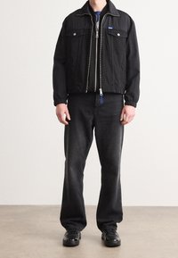 Una persona che indossa una giacca nera con zip, jeans scuri e sneakers nere, in piedi su un pavimento grigio con uno sfondo chiaro e semplice.