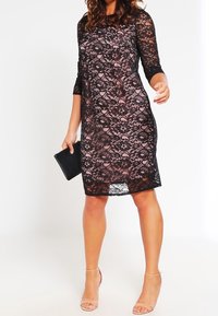 Abito in pizzo nero con maniche a tre quarti, motivo floreale trasparente su una fodera color nudo, taglio dritto, lunghezza al ginocchio, abbinato a una clutch nera.