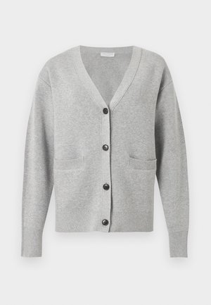 Naine, kes kannab halli nööpidega kardigaani, laia säärega valgeid pükse ja musti teravate ninadega slingback-kingi, seisab tavalise tausta ees.