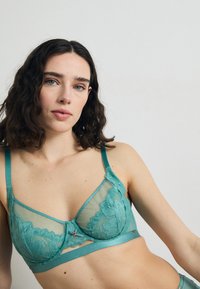 Mėlyna nėrinių braletė su permatomais elementais, gėlių raštais, reguliuojamomis petnešėlėmis ir dekoratyviu priekyje esančiu užsegimu. Lygus satino apatinis kraštas.