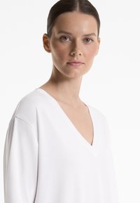 Haut blanc en V, fabriqué en tissu lisse, avec des épaules tombantes et une coupe décontractée. Design simple et épuré sans motifs visibles.