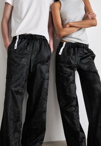 Black, glossy fabric cargo pants with elastic waistband and drawstring. Features side pockets and contrasting tags; textile appears smooth.