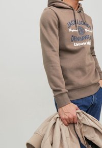 Sudadera marrón con estampado de logo, tela suave, bolsillo canguro, puños acanalados y dobladillo. Se sostiene en la mano una chaqueta beige, también texturizada.