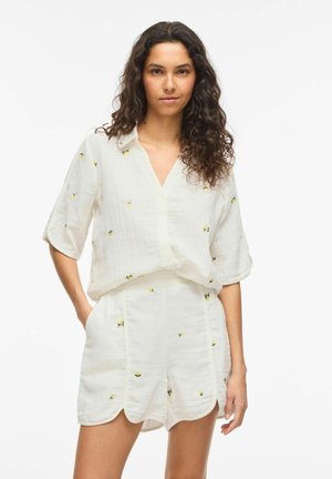 Kvinne med langt, krøllete hår iført hvit kortermet skjorte og matchende scalloped shorts med små gule blomstertrykk.
