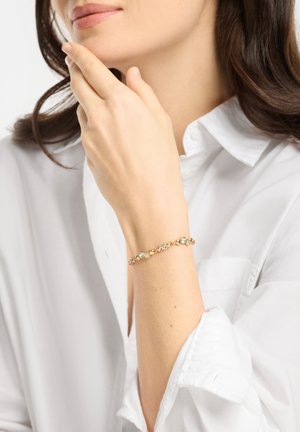 Valkoista paitaa käyttävä nainen koskettaa leukaa ja näyttää ranteessaan kultaisen rannekorun, jossa on monivärisiä jalokiviä.