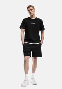 Schwarzes T-Shirt mit dem Aufdruck "LAZY CLUB" in Weiß, kombiniert mit schwarzen Shorts und weißen Sneakern. Das Modell steht mit den Händen in den Taschen vor einem neutralen Hintergrund.