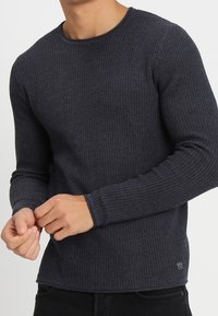 Ciemnoniebieski sweter z długim rękawem o teksturowanej powierzchni z okrągłym dekoltem i dopasowanymi rękawami. Posiada widoczne szwy oraz małe logo na hemie.