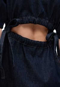 Κοντινό πλάνο σκούρου μπλε denim outfit με ελαστική μέση που σφίγγει και δέσιμο στο πλάι, αποκαλύπτοντας τμήμα γυμνού μέσης.